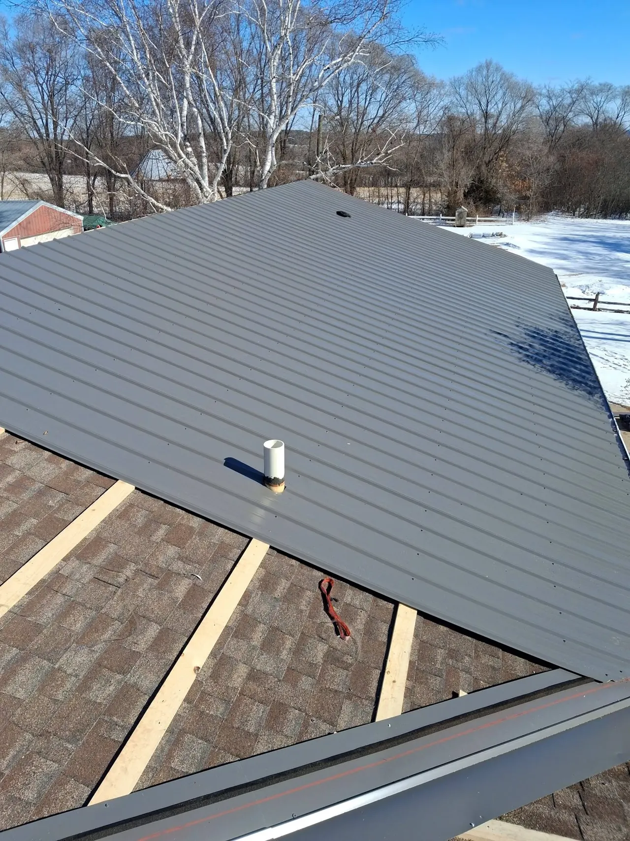 Gray ribbed metal roof panels beside exposed underlayment and timber strips, with snow and trees beyond.