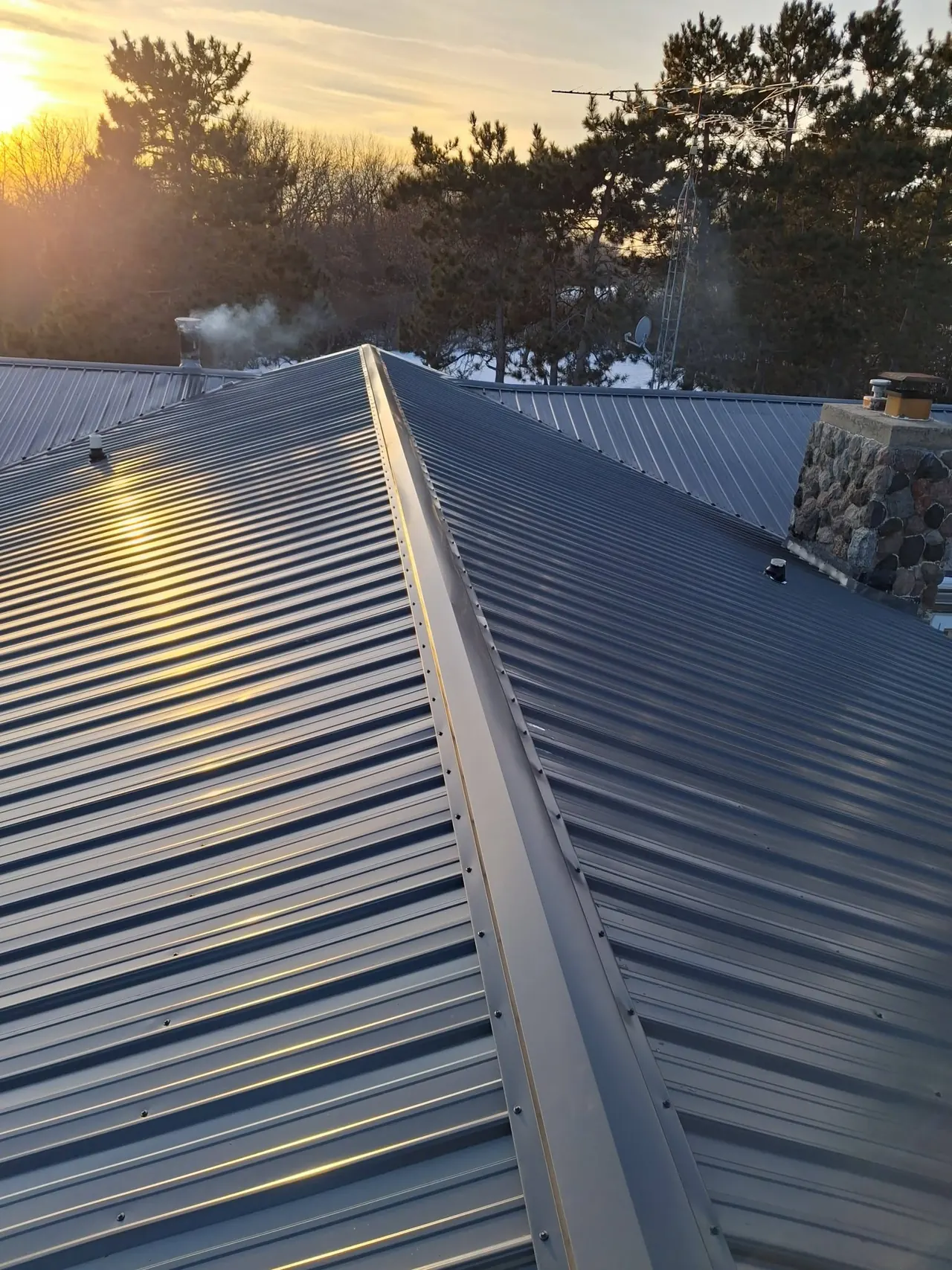 Gray ribbed metal roof ridge in low sunlight, with a stone chimney and trees beyond.