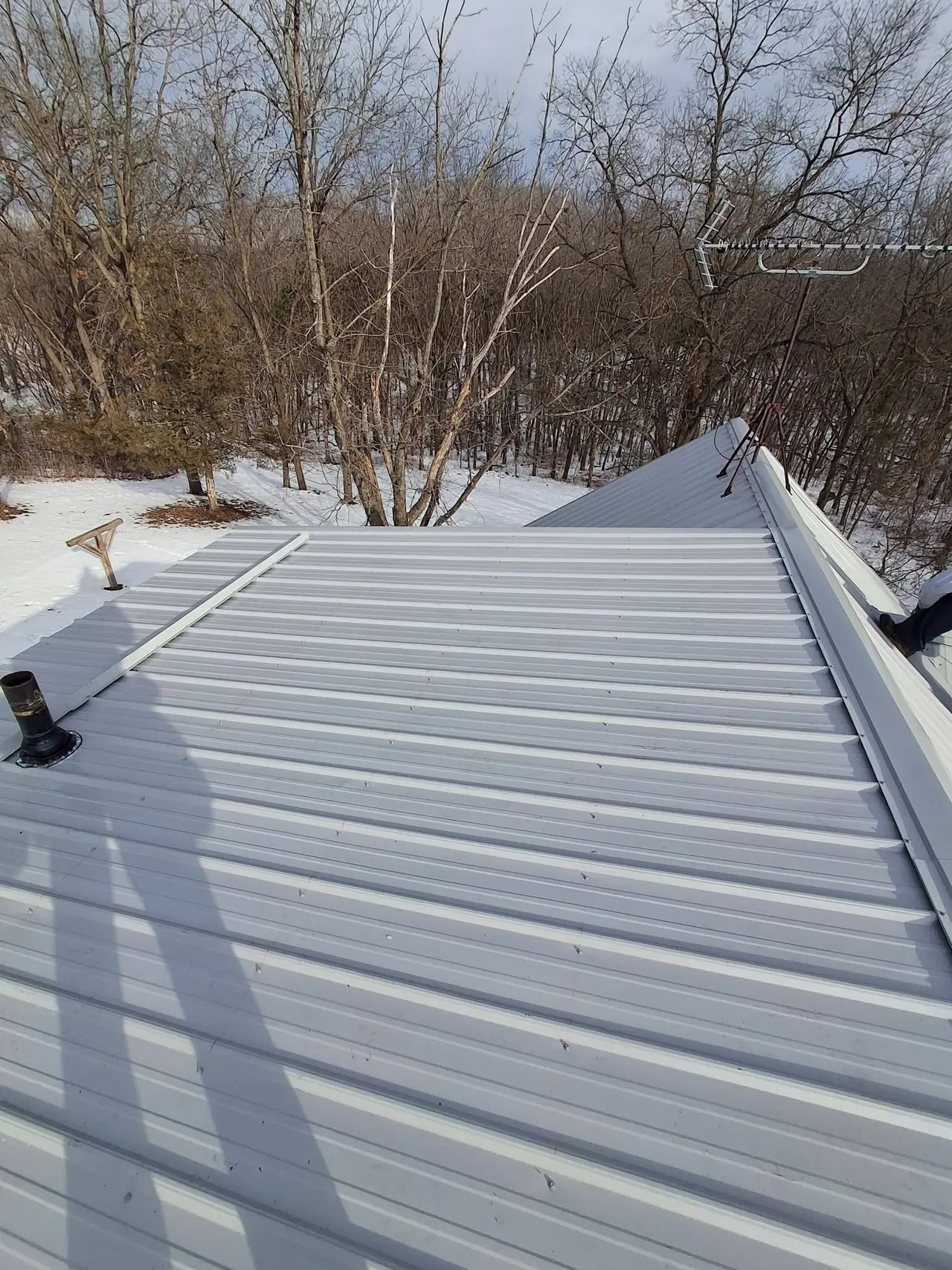 Gray metal roof slope and ridge beside an antenna, with snow and bare trees beyond.